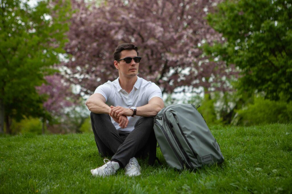 A young man in a white shirt and sunglasses sits on the grass, leaning against his backpack, near cherry blossom trees. 