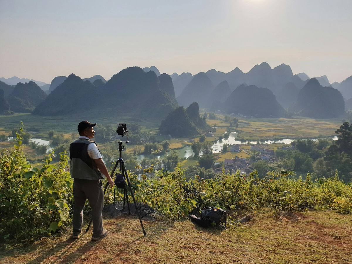 Man standing on top of a mountain taking a photo with camera on a tripod