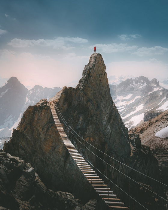 Man on tall rocky peak with rope bridge over valley